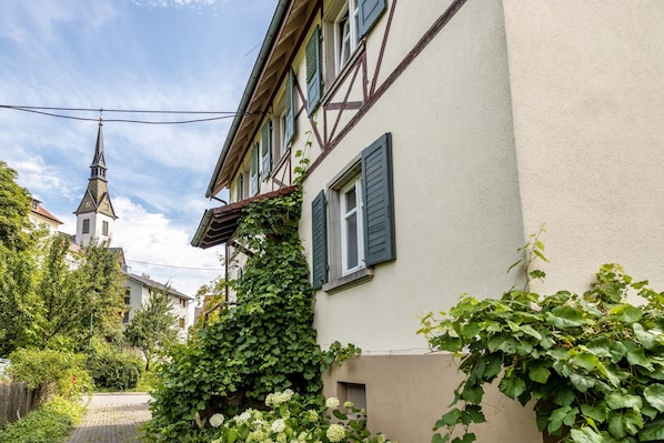 A narrow street with a church steeple, a building with green shutters, and lush greenery.