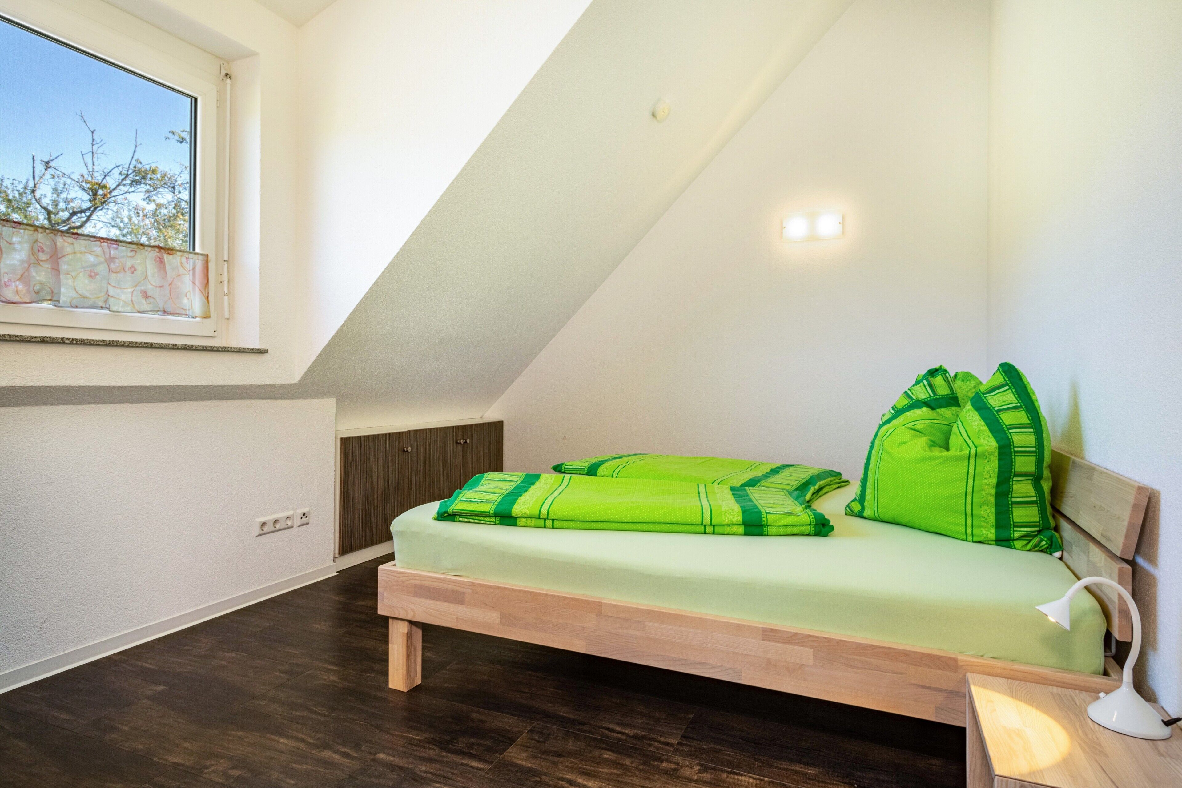 Dormitorio tipo loft con techo inclinado, cama con ropa de cama verde, lámpara de noche y ventana con vista a árboles.