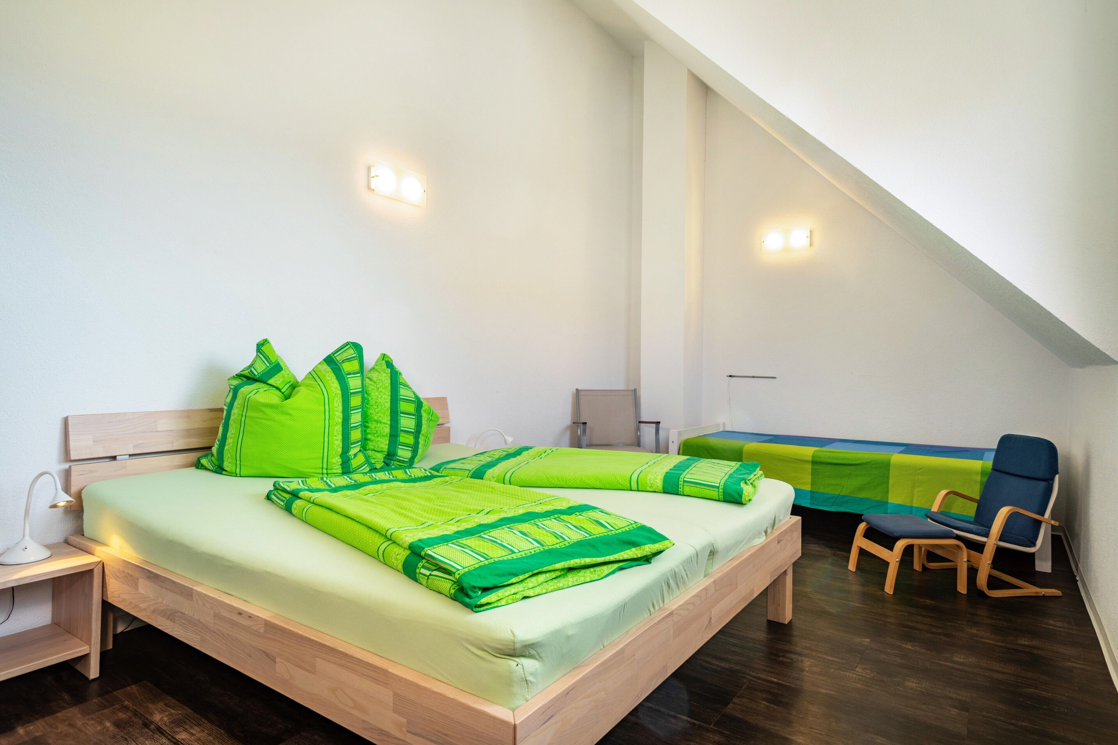 Ein Schlafzimmer mit einem hölzernen Bett, grüner Bettwäsche, einer Bank, einem Sessel und einem Schreibtisch mit Lampe.