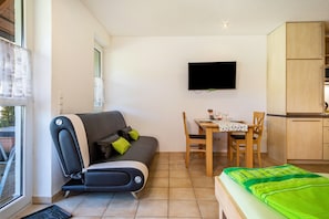 Ein kompakter Raum mit Sofa, Esstisch, Wandfernseher und Bett mit grüner Decke.