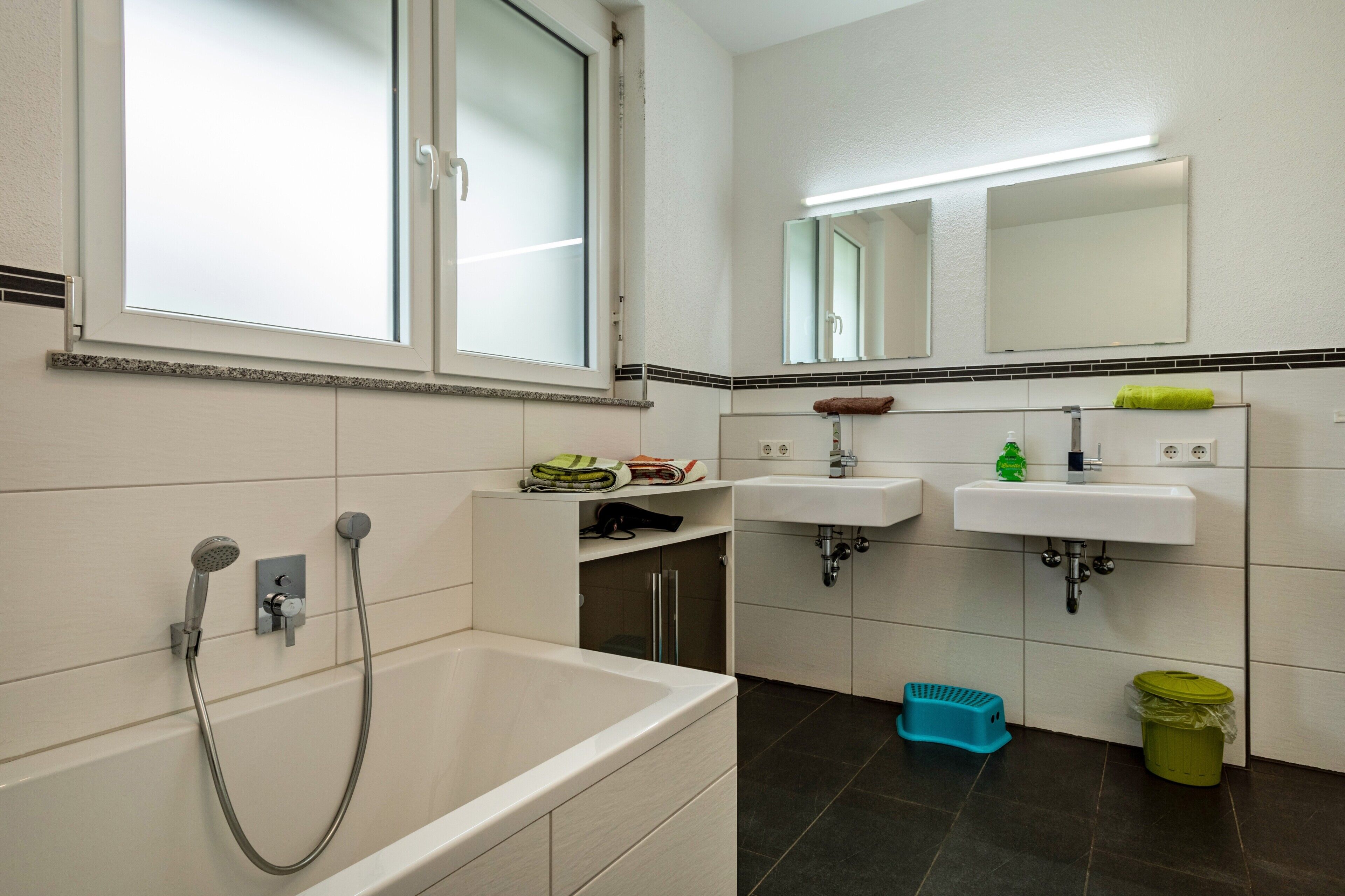 Een moderne badkamer met een badkuip, twee wastafels, een raam en een spiegel.
