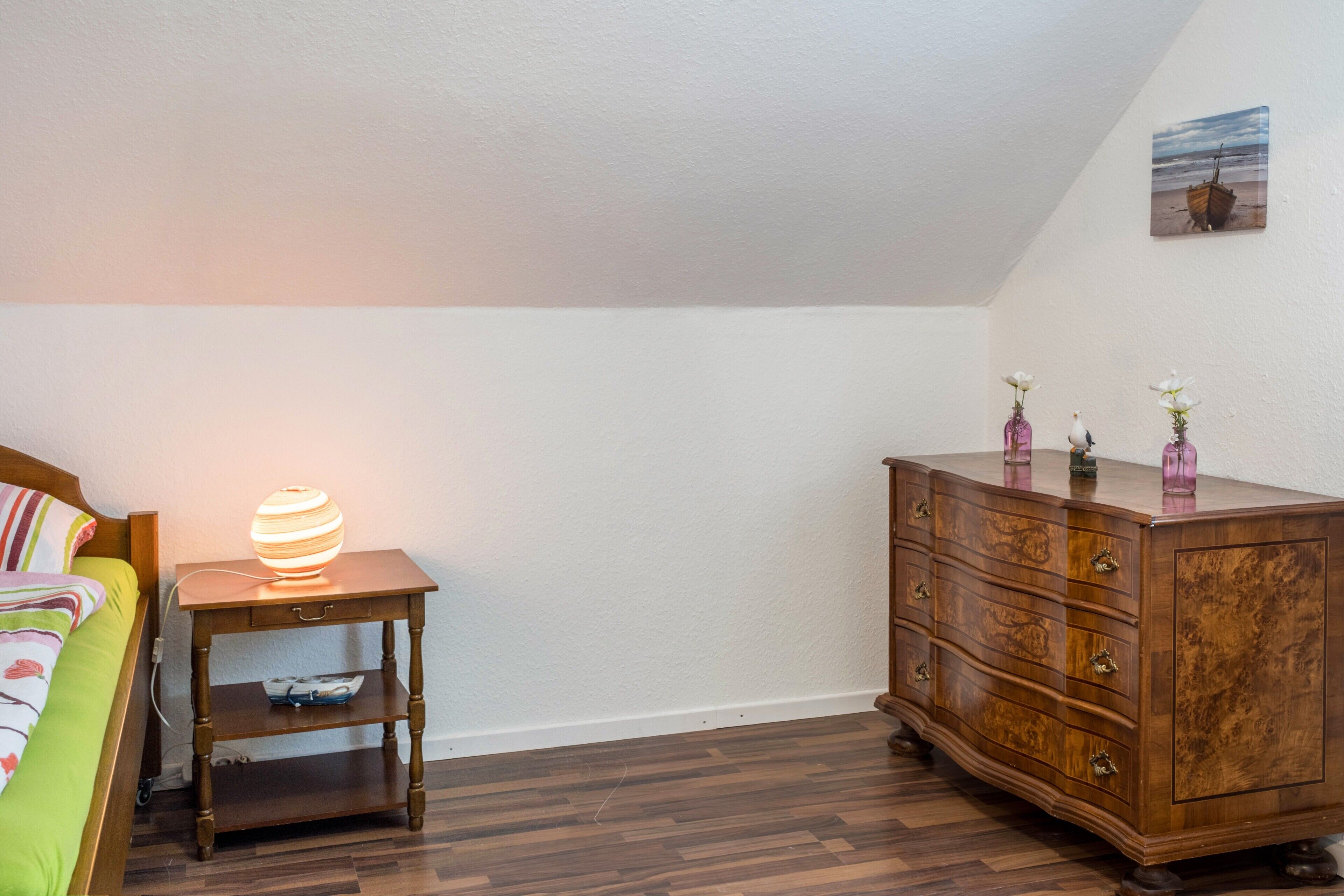 A bedroom with a bed, a nightstand with a lamp, a dresser, and a painting on the wall.