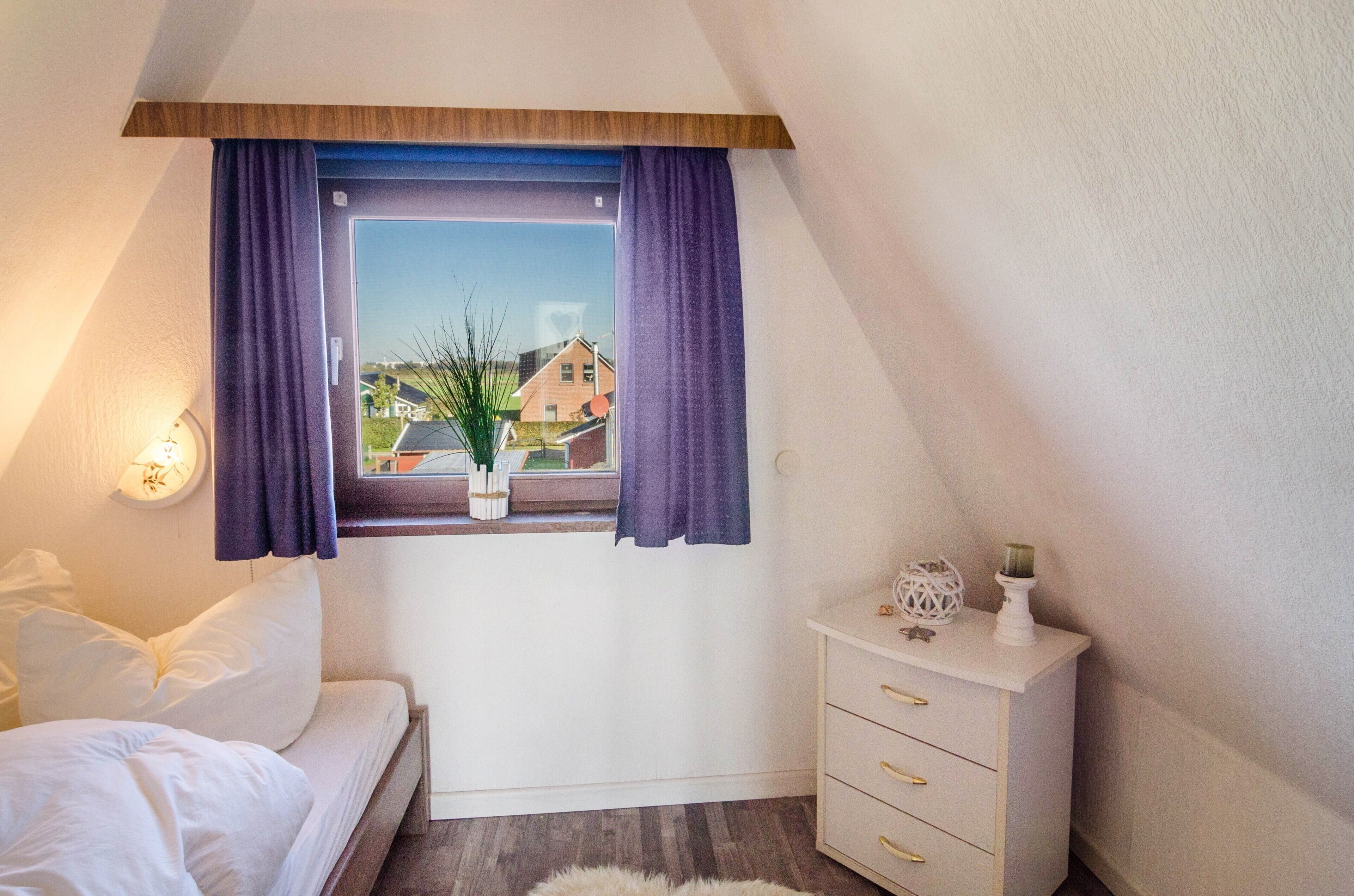 A bedroom with a bed, a window with curtains, a bedside table with a lamp, and a view of houses outside.