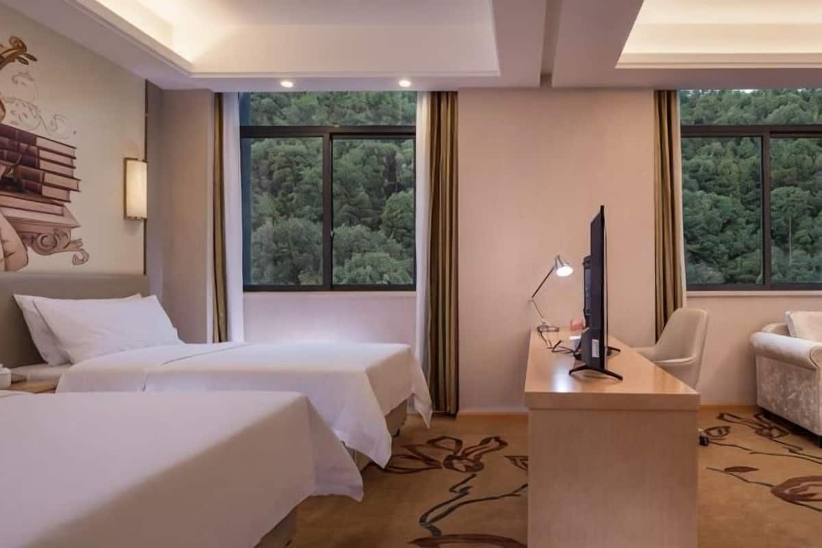 Habitación de hotel con dos camas, un escritorio con lámpara, una silla y vistas a árboles a través de amplias ventanas.