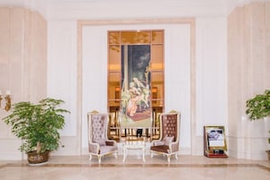 A lobby with a reception desk, two chairs, a large painting, and potted plants.