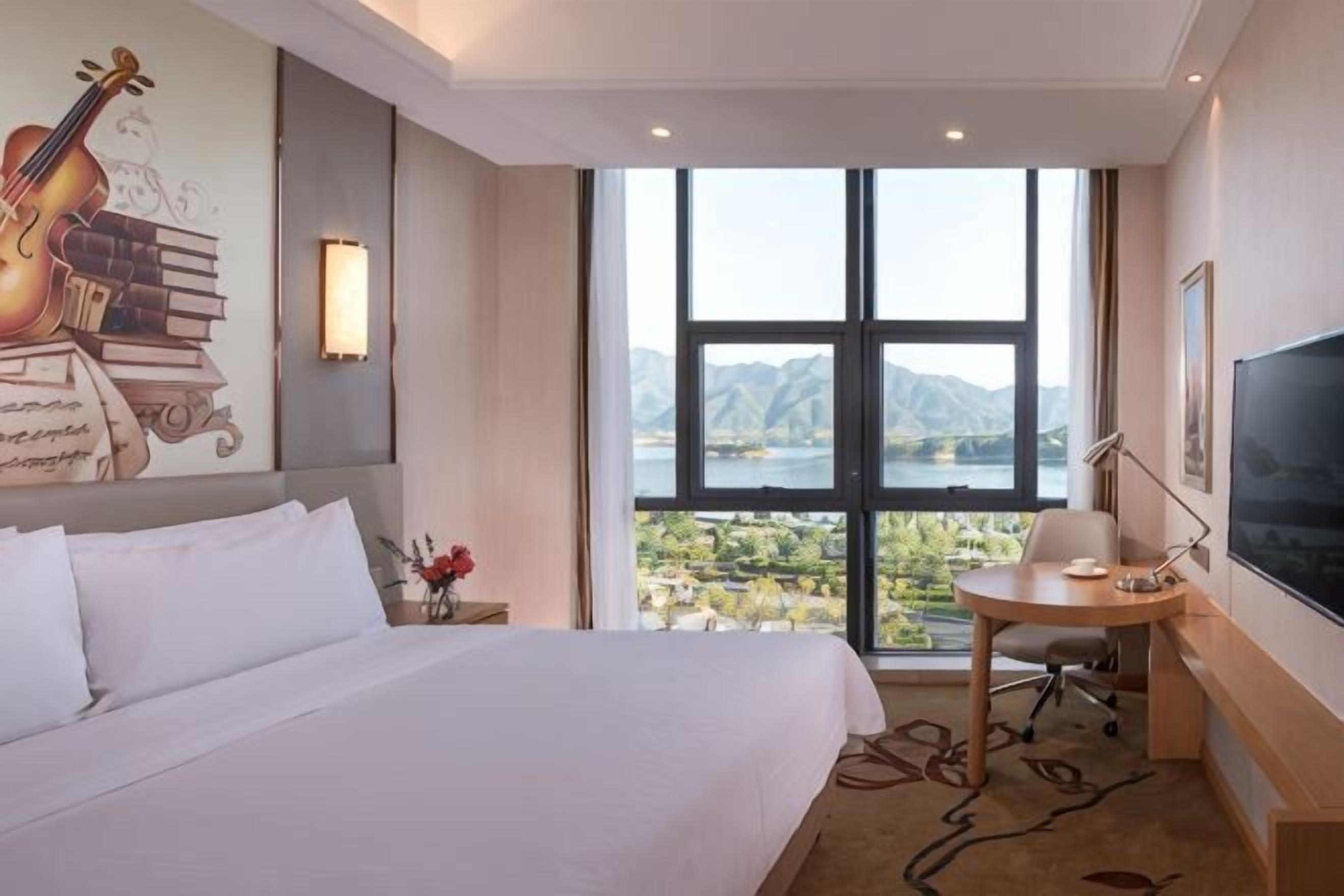 A hotel room with a large bed, a desk with a chair, a TV, and a view of mountains through a large window.