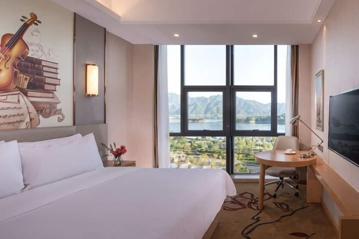 Habitación de hotel con una cama grande, un escritorio con silla, televisión y vistas a montañas a través de un ventanal amplio.