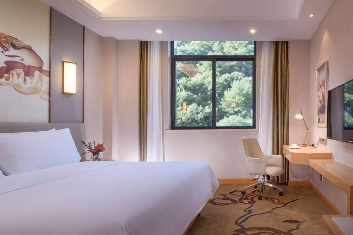 Habitación de hotel con cama, escritorio, silla, ventana con vista a árboles y un cuadro en la pared.