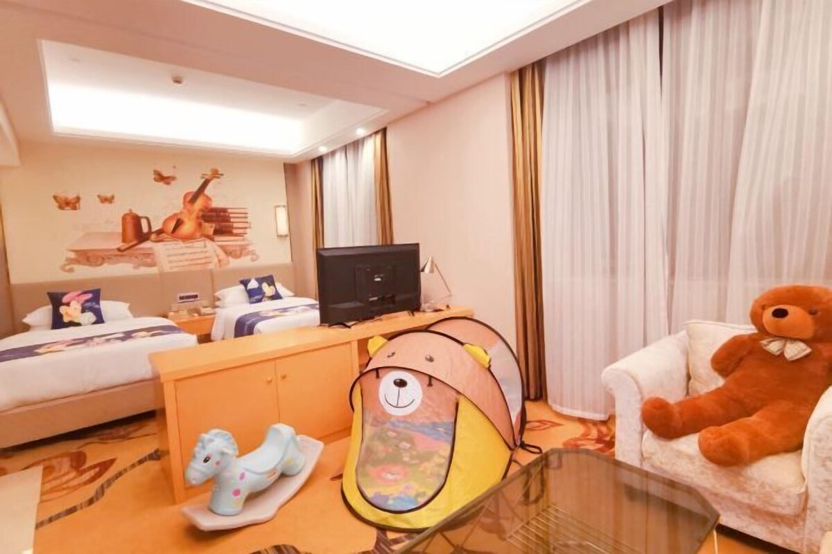 Una habitación de hotel con cama, un escritorio con computadora, un osito de peluche, una tienda de campaña infantil y una mesa de vidrio.