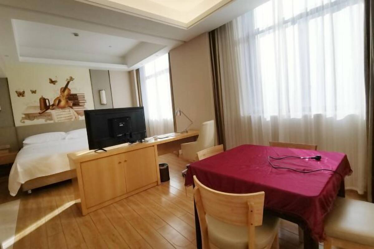Habitación de hotel con cama, escritorio con computadora, mesa con mantel rojo y ventana con cortinas.