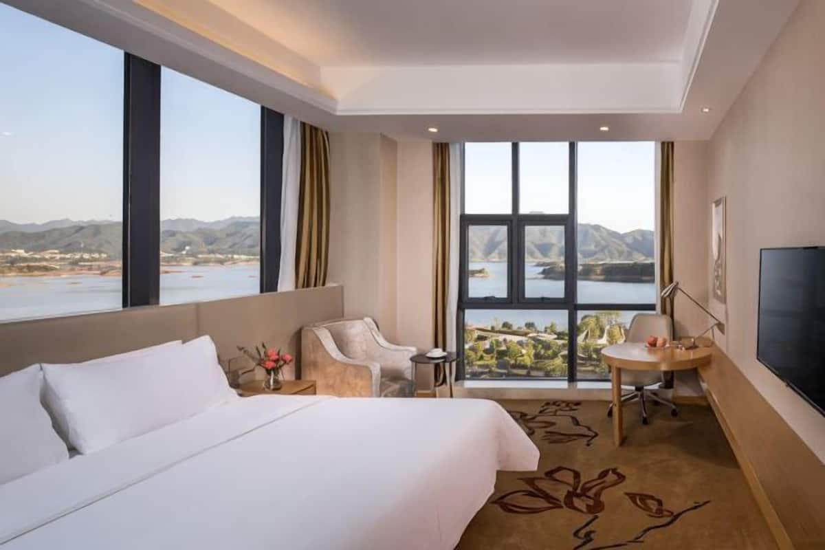 Habitación de hotel con una cama grande, un escritorio, una silla, un televisor y vistas a un lago y montañas.