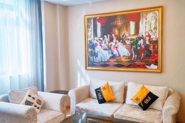 Una habitación con un cuadro clásico grande en la pared, un sofá con almohadones decorativos y una ventana con cortinas translúcidas.