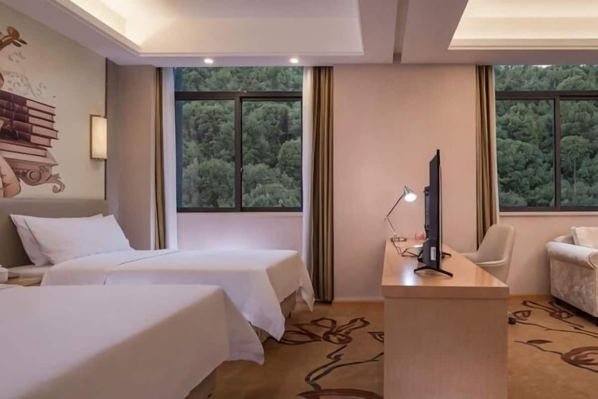 Habitación de hotel con dos camas, un escritorio con lámpara, una silla y vistas a árboles a través de la ventana.