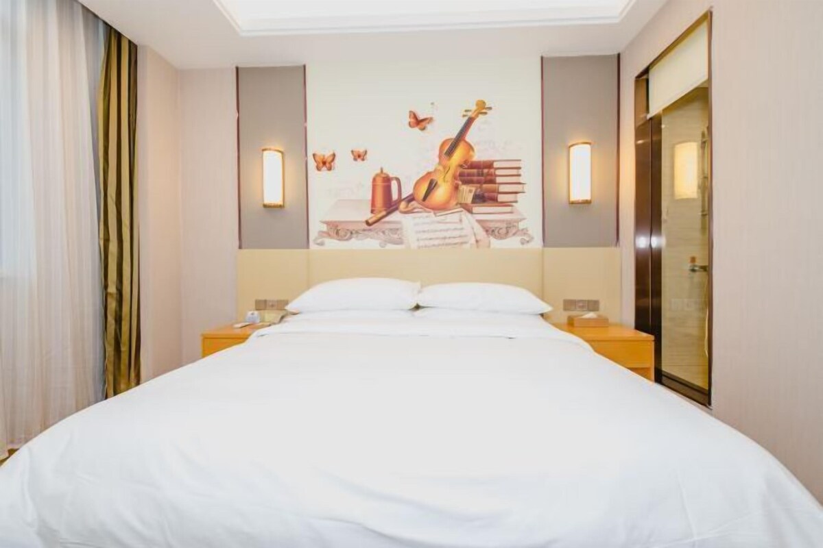Habitación de hotel con cama, mesita de noche, lámpara de pared y un cuadro grande de un violín y libros.