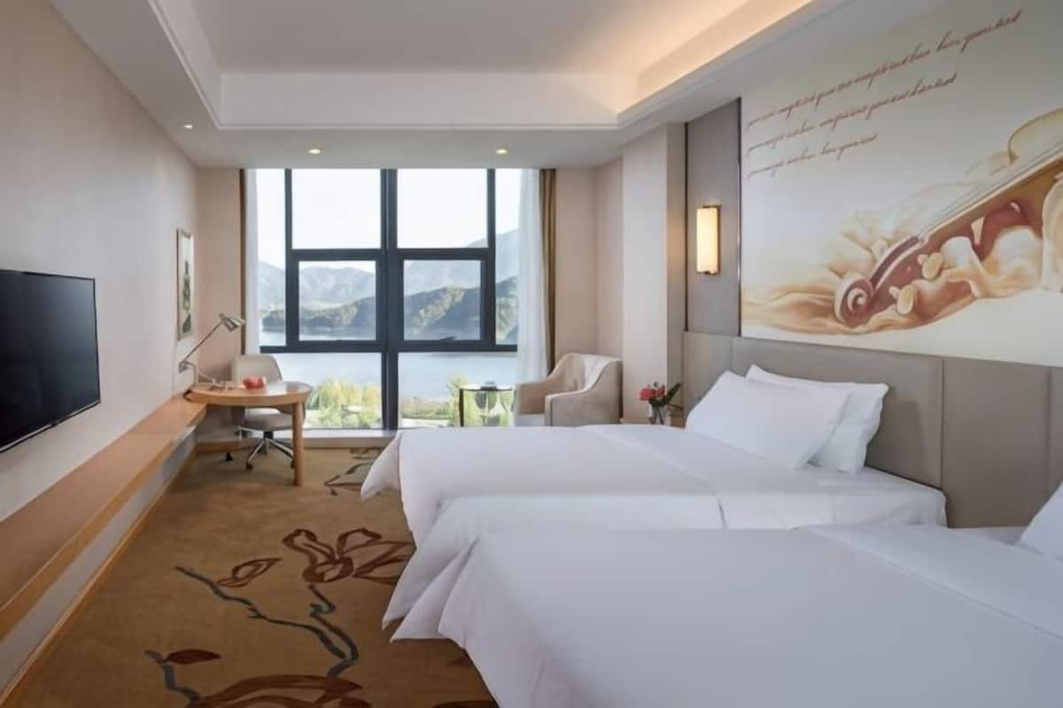 Habitación de hotel con dos camas, televisión, escritorio, silla, ventana con vistas y un adorno en la pared.