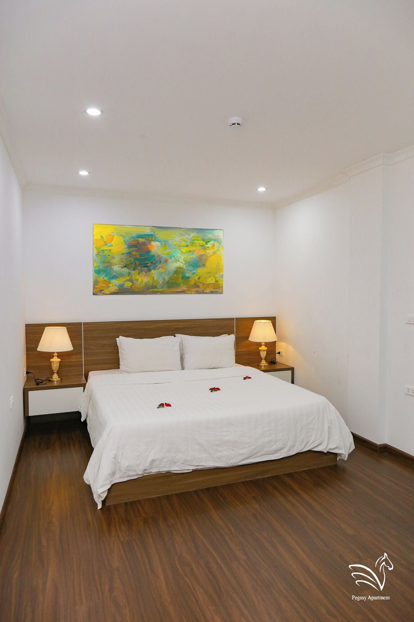 Une chambre d’hôtel avec un lit en bois, du linge de lit blanc, deux lampes de chevet, un luminaire au plafond et une peinture abstraite aux couleurs vives accrochée au mur.