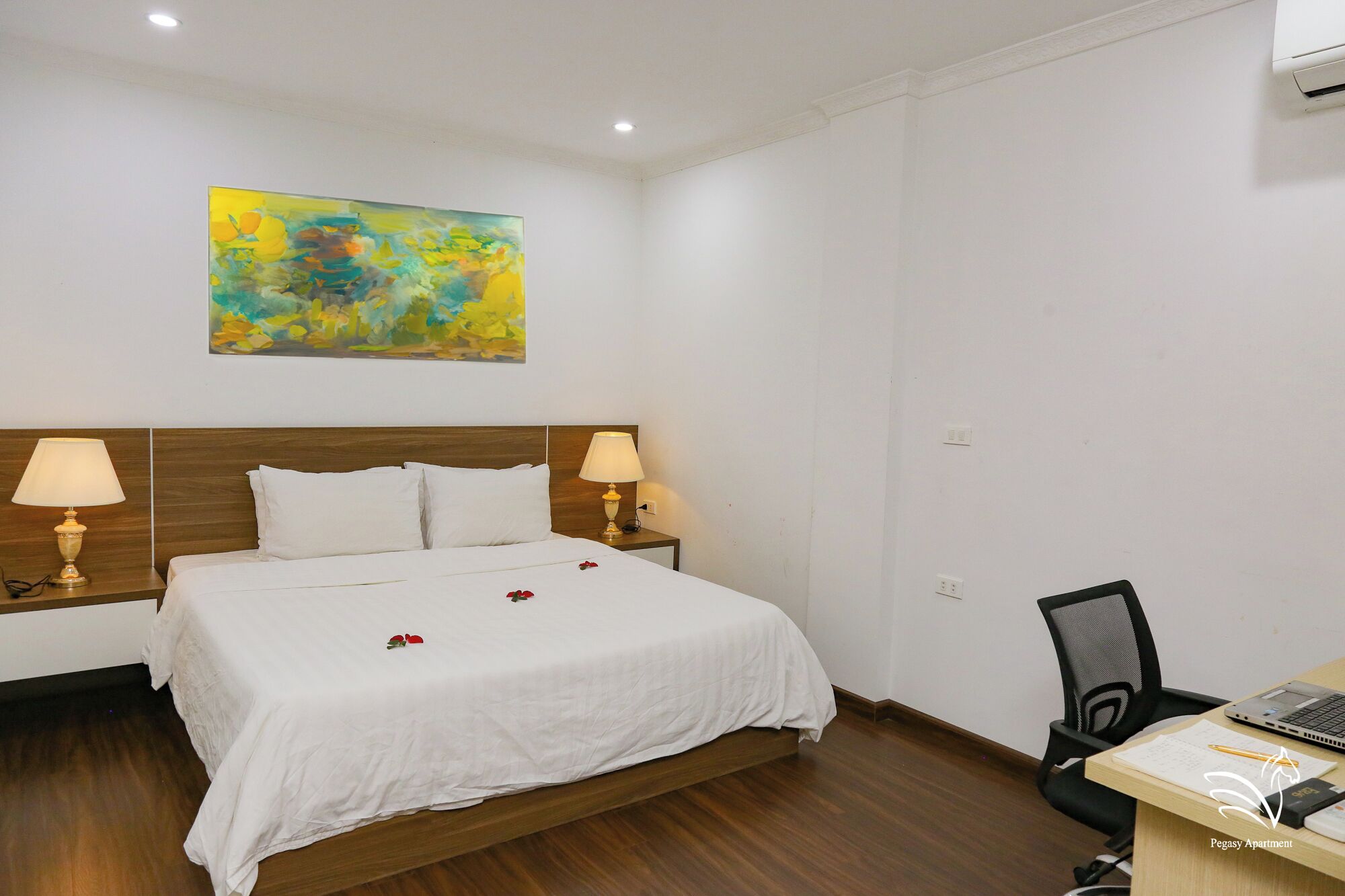 A hotel room with a bed, two bedside lamps, a desk with a chair, and a painting on the wall.