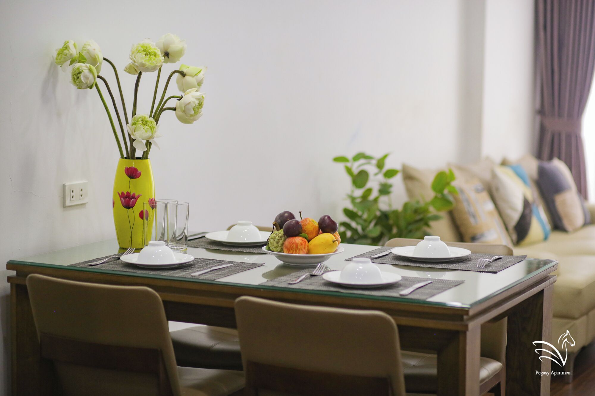 A dining table set with plates, cutlery, and a bowl of fruit. A vase with flowers is on the table.