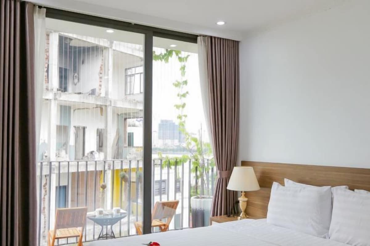Une chambre d’hôtel avec un lit, une table de chevet, une lampe, une fenêtre avec des rideaux et une vue sur les bâtiments environnants.