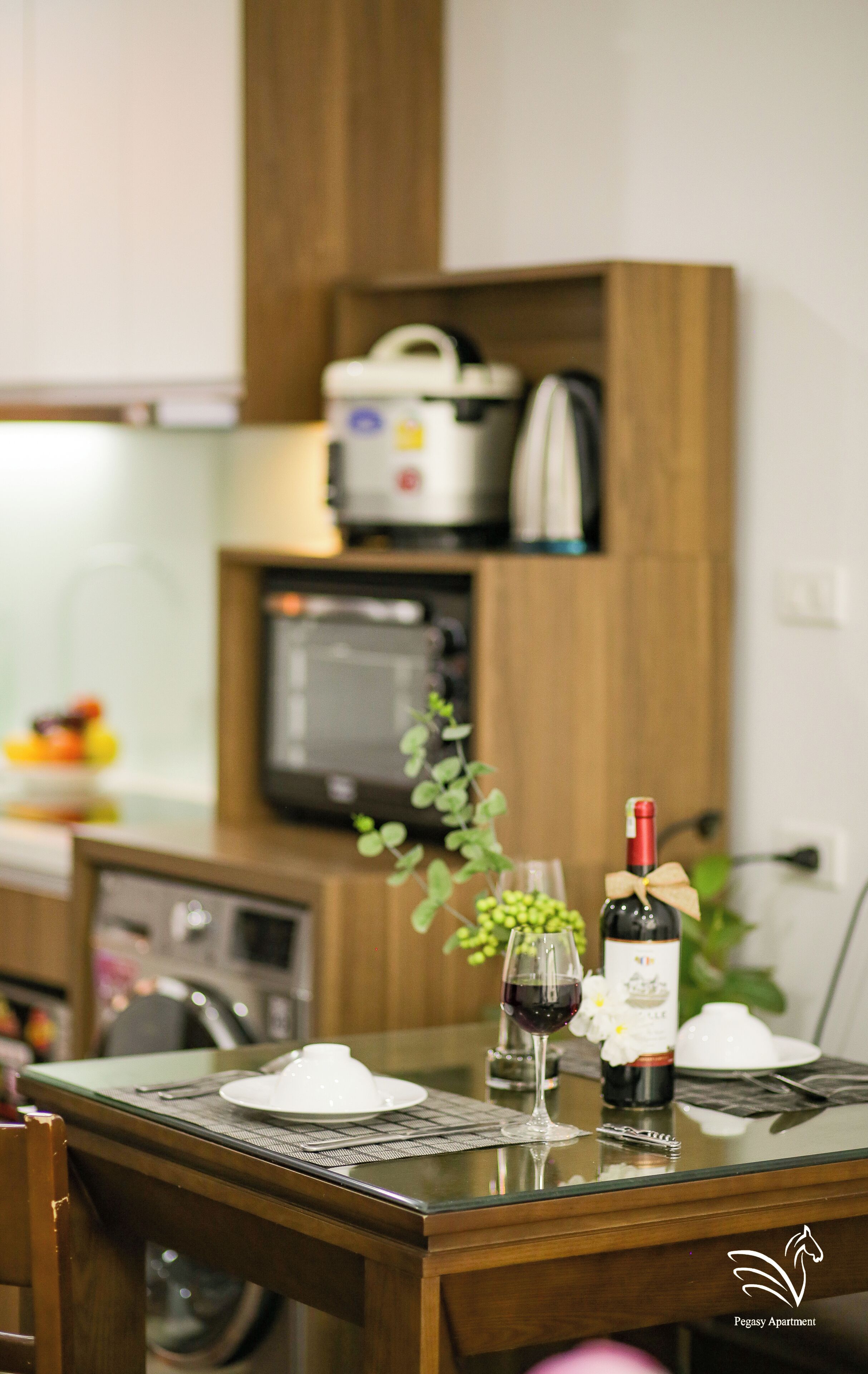 Une table à manger avec une bouteille de vin, un verre de vin et une petite plante. On aperçoit un four à micro-ondes et un cuiseur à riz à l’arrière-plan.