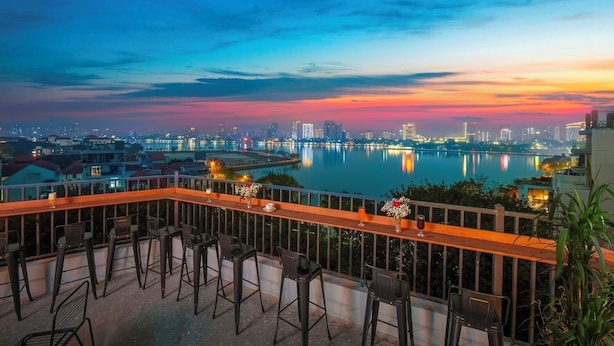 A terrace with bar stools, a railing, and a view of a city skyline at dusk. - Pegasy Apartments & Travel (Hanoi)