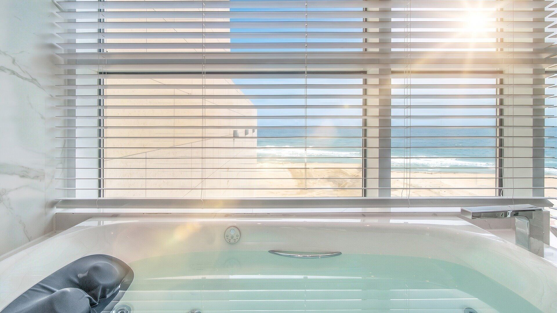 A bathroom with a bathtub, a window with blinds, and a view of the beach.