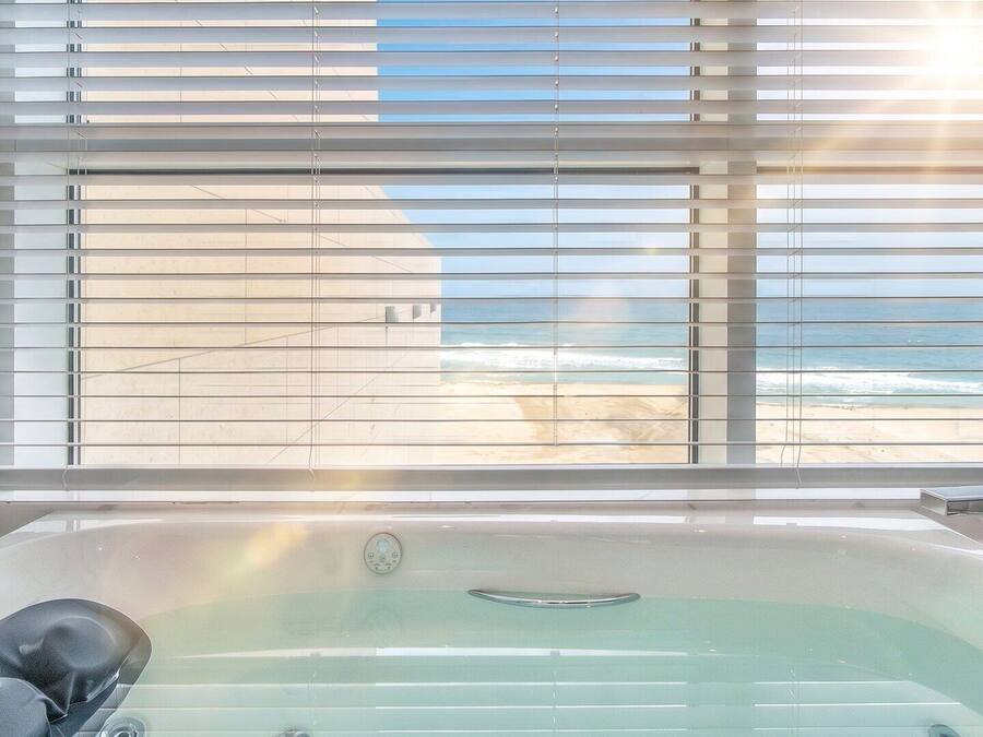 A bathroom with a bathtub, a window with blinds, and a view of the beach.