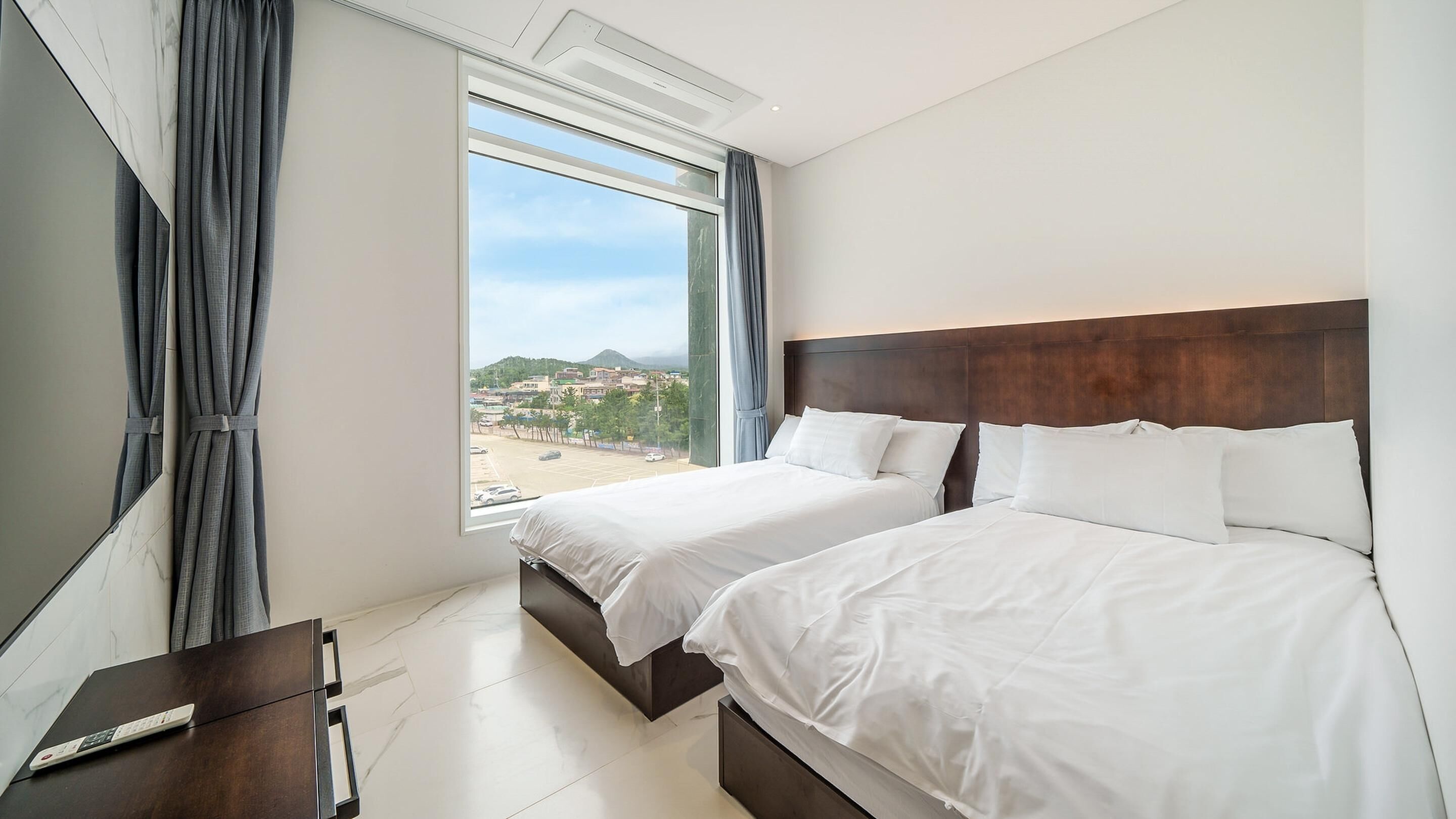 A hotel room with two beds, a large window with curtains, a flat-screen TV, and a remote control on a wooden table.