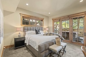 A bedroom with a large bed, a nightstand, a bench, and a view of the outdoors through a sliding glass door.