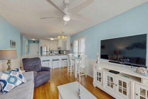 A living room with a sofa, a TV, a ceiling fan, and a kitchen in the background.