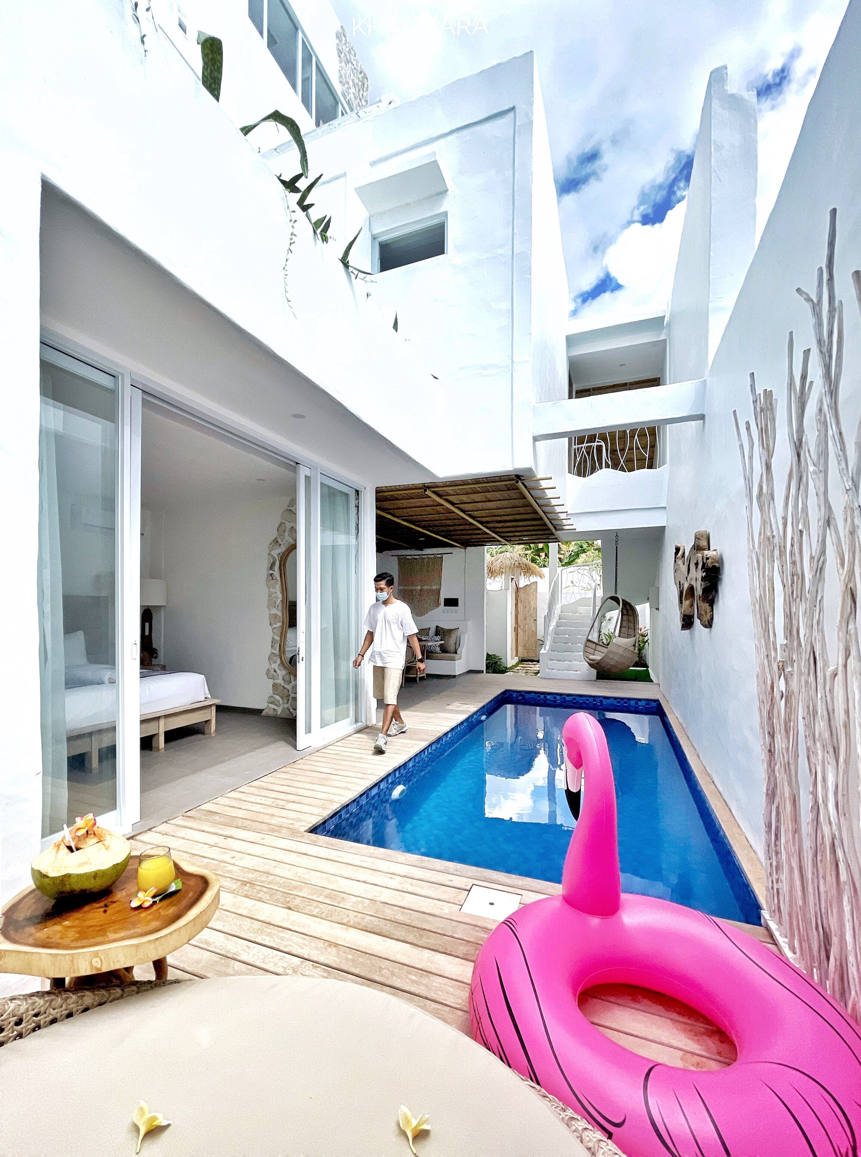 A modern outdoor pool area with a wooden deck, a pink inflatable ring, and a bed with a white canopy.