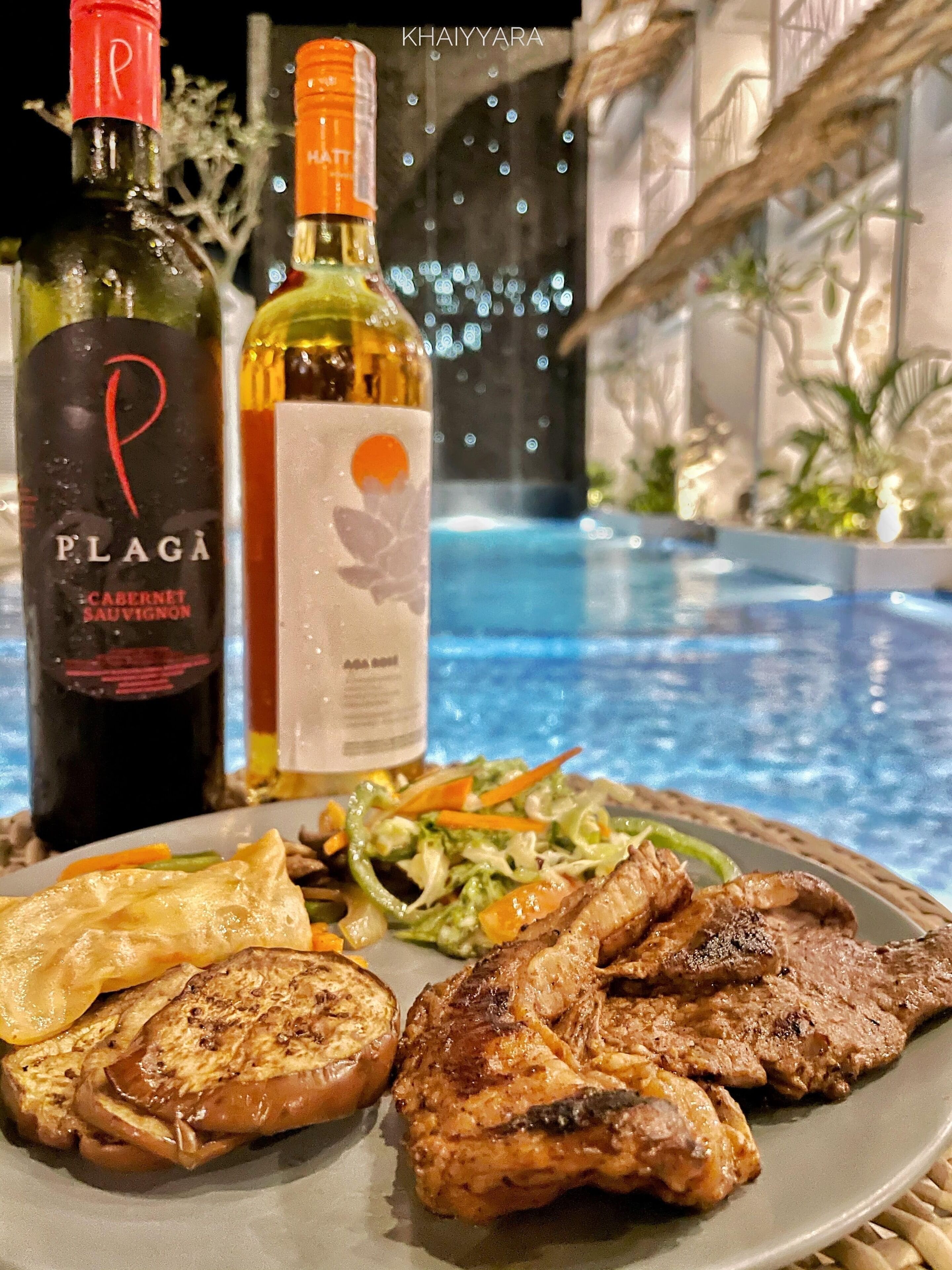 A dining table with a plate of grilled meat, vegetables, and toast, and two bottles of wine.