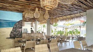 Ein Restaurant im Strandhausstil mit Korbmöbeln, hängenden Lampen und einer Wandmalerei mit Strandmotiv.