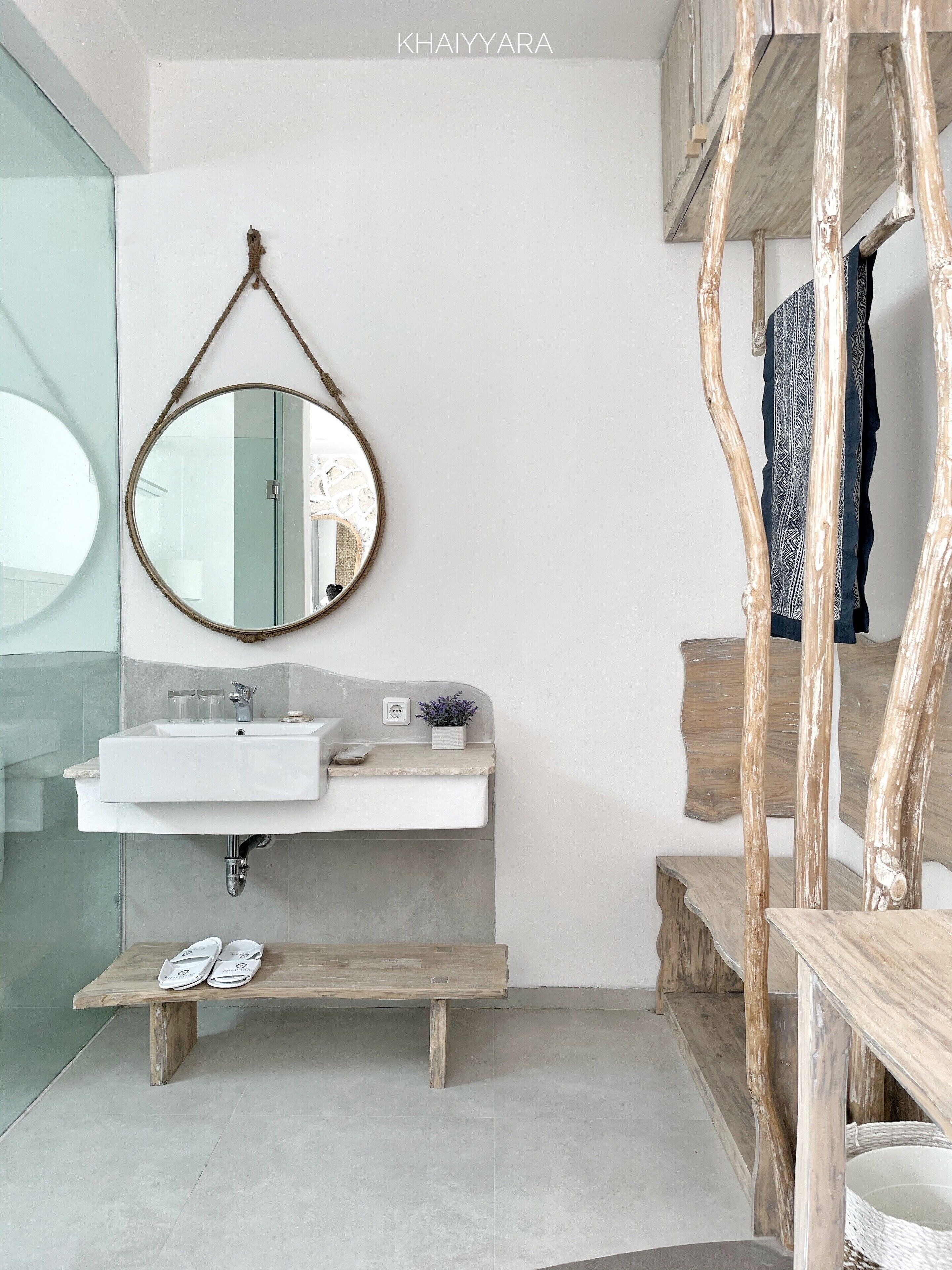 Un baño moderno con un espejo redondo, lavamanos blanco y banco de madera.