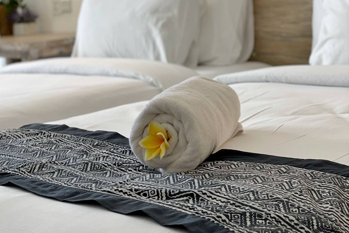 Ein ordentlich bezogenes Bett mit weißen Leinen, ein aufgerolltes Handtuch mit einer gelben Blume und eine dekorative Decke.