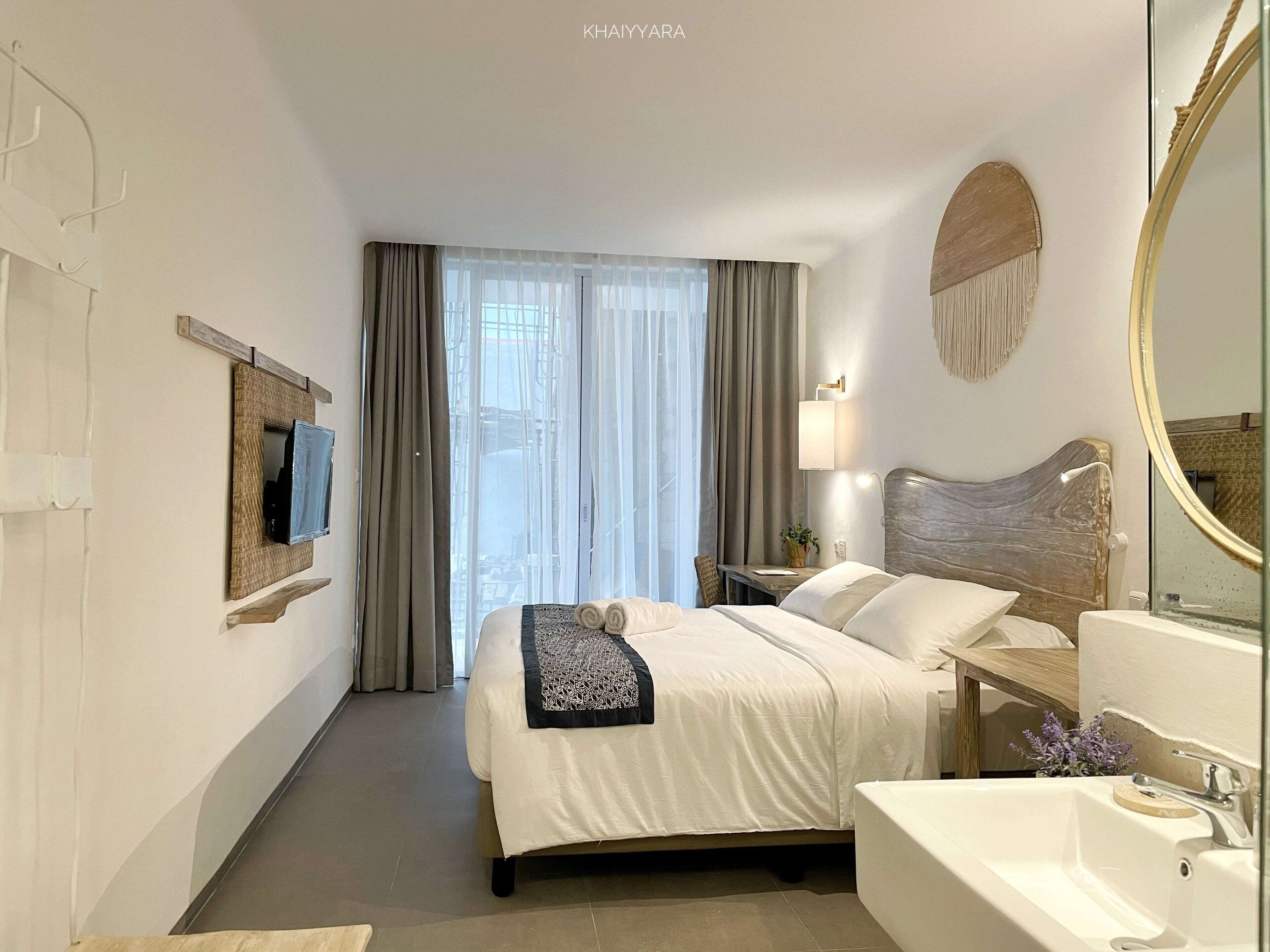 A hotel room with a bed, a bedside table with a lamp, a large mirror, a TV mounted on the wall, and a window with curtains.