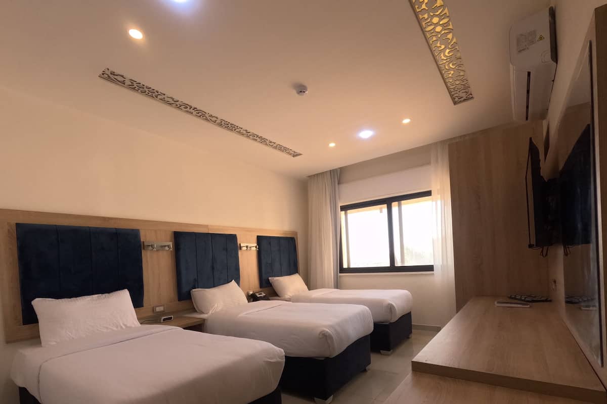 A hotel room with three beds, a wooden desk, a large window, and a flat-screen TV.