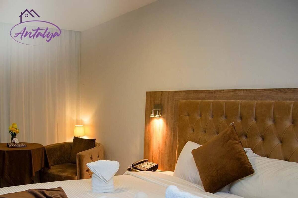 A hotel room with a large bed, a headboard, a bedside table with a lamp, a chair, and a small table with a vase of flowers.