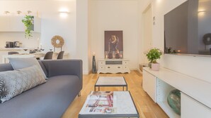 Un salon moderne avec un canapé gris, une table basse sur laquelle sont posés des magazines, un coin repas avec une table dressée pour deux personnes, et une cuisine en arrière-plan.