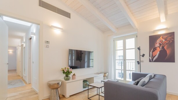 Un salon moderne avec un canapé gris, un téléviseur à écran plat, une petite table basse et un vase décoratif. Une porte donne accès à un balcon avec vue sur l’extérieur.