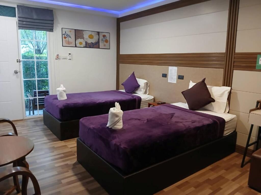 A hotel room with two beds, each with a purple bedspread and white pillows. There is a wooden floor, a small table, and a window with a view of greenery outside.