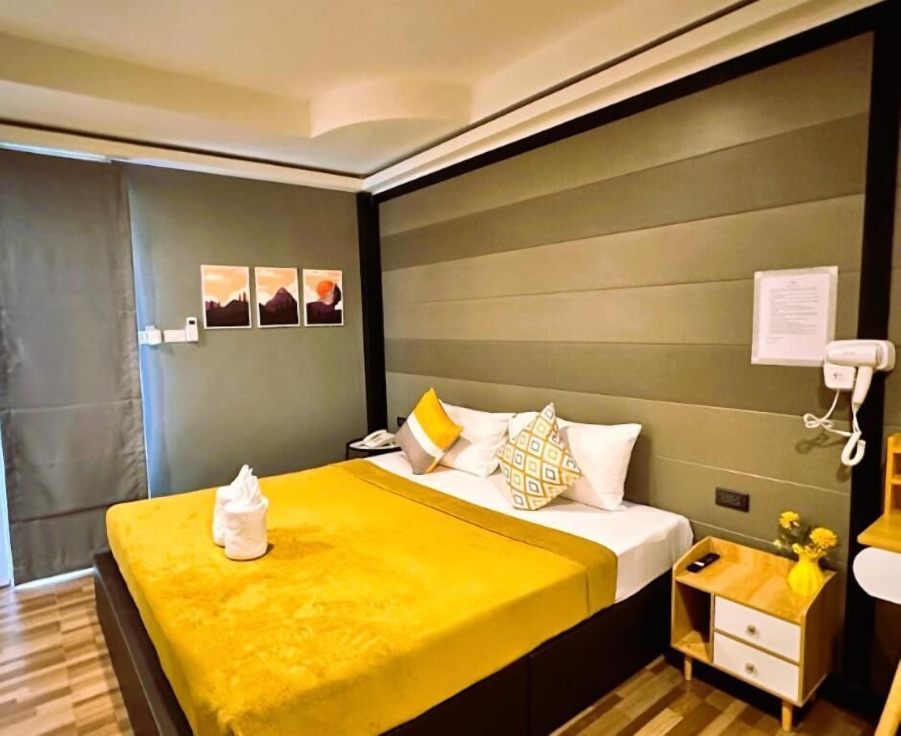 Een hotelkamer met een bed, nachtkastje en een bijzettafeltje met een telefoon en een bloevas.