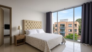 A modern bedroom with a large bed, a nightstand, a lamp, and a view of buildings through large windows.