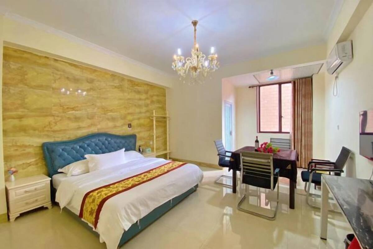 A bedroom with a bed, a desk, a chair, a table, a chandelier, and a window.