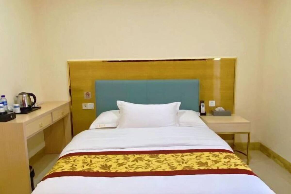 A hotel room with a bed, a bedside table with a lamp, a small desk with a kettle, and a wall-mounted phone.