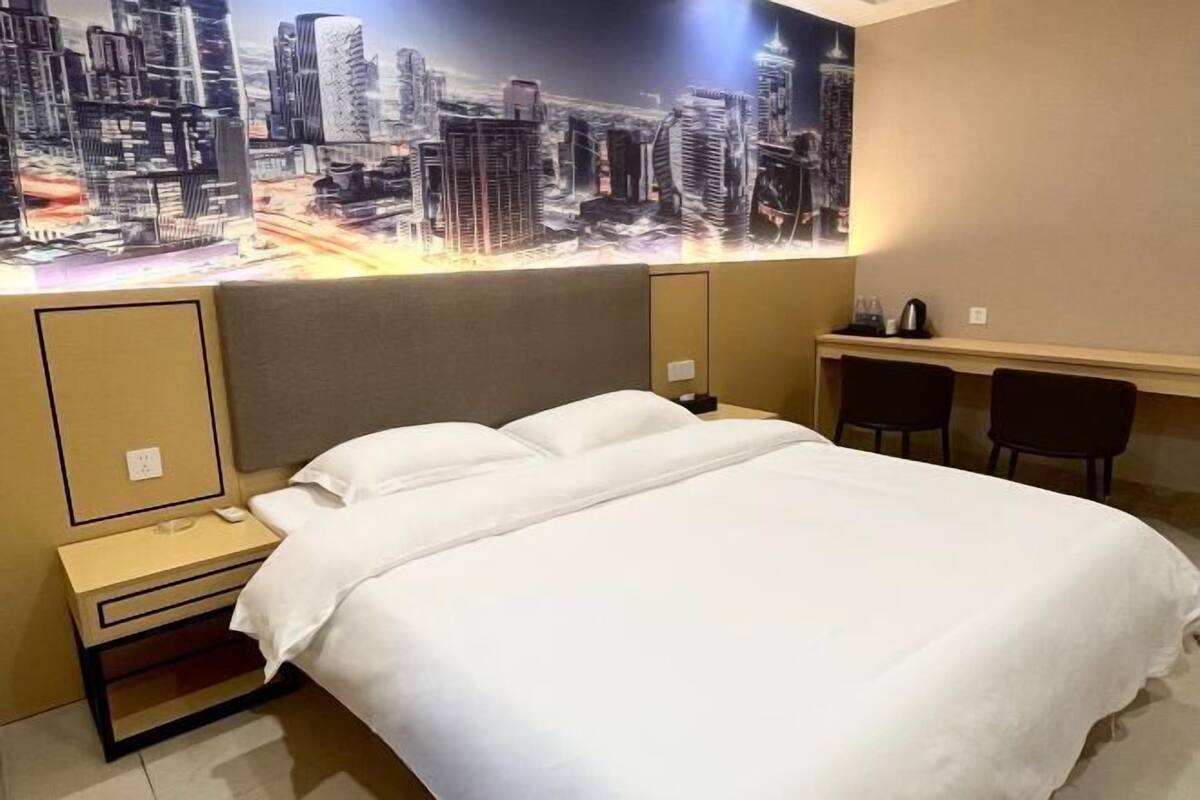 A hotel room with a large bed, a nightstand, a desk with two chairs, and a cityscape mural on the wall.