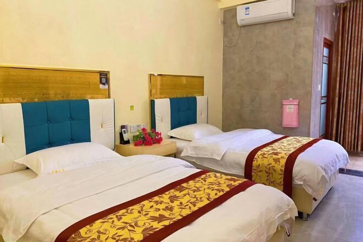 A hotel room with two beds, each with a headboard, a bedside table with a lamp, and a wall-mounted air conditioner.