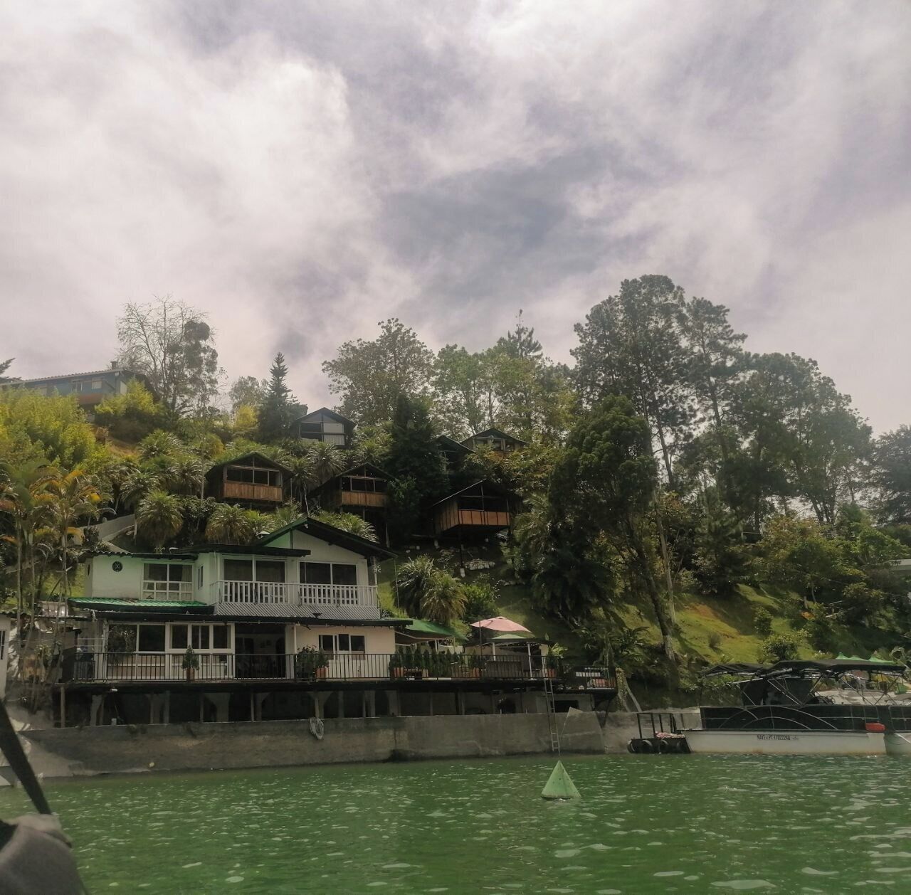 A coastal property with multiple buildings, including a main house with a balcony and a smaller structure, surrounded by lush greenery and overlooking a body of water.