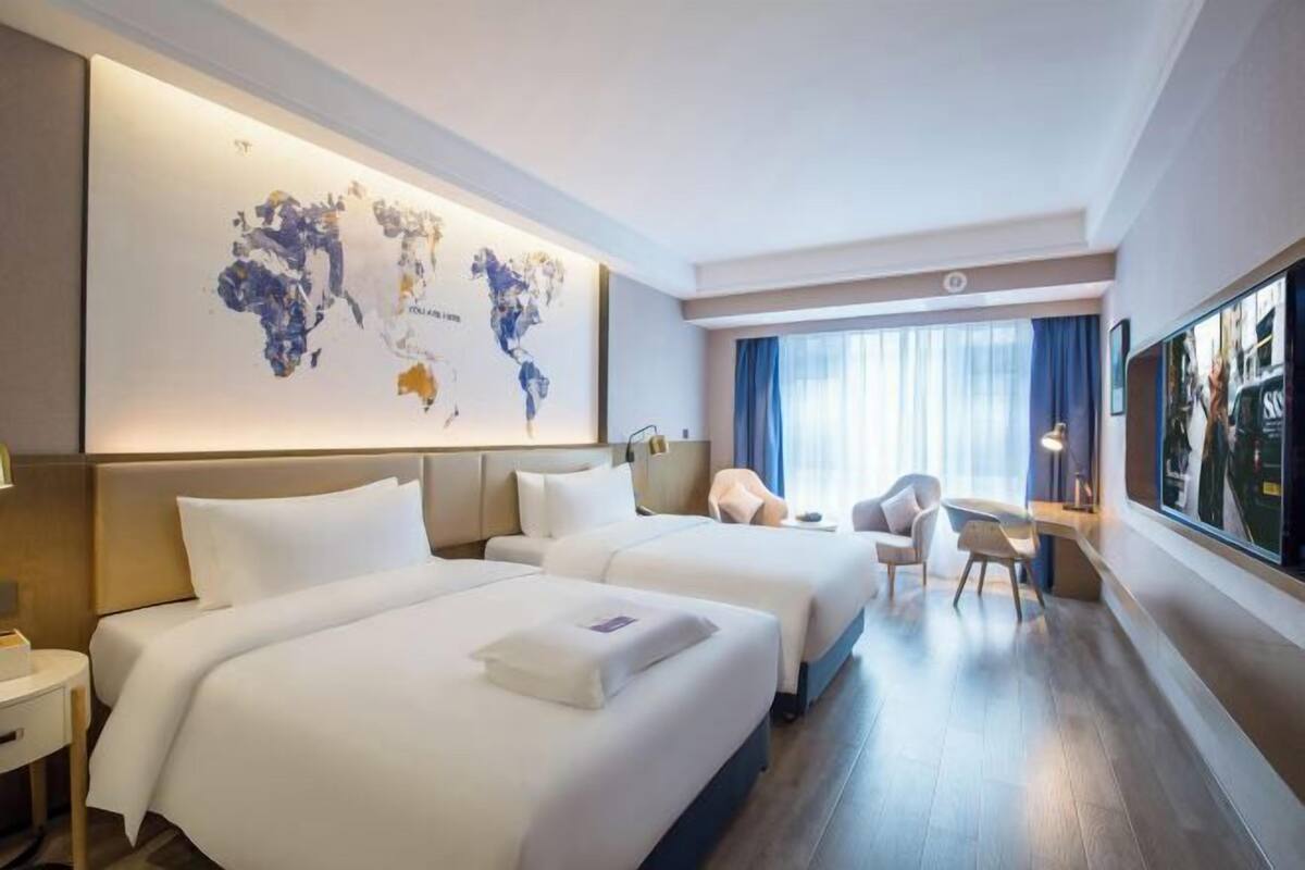 A hotel room with two beds, a large world map on the wall, a desk with a chair, and a large window with curtains.