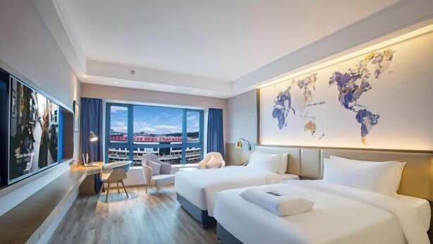 A hotel room with two beds, a desk, a chair, a large window with a view, and a world map on the wall. - Guiyi Hotel (Guiyang Guanshanhu District Southwest International Trade City Store) (Guiyang)